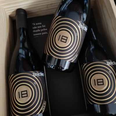 Três garrafas de vinho com rótulos pretos e dourados dentro de caixa de madeira clara