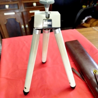 Tripé de câmara metálico prateado com punho cinza e etiqueta Universal Made in Germany sobre mesa vermelha