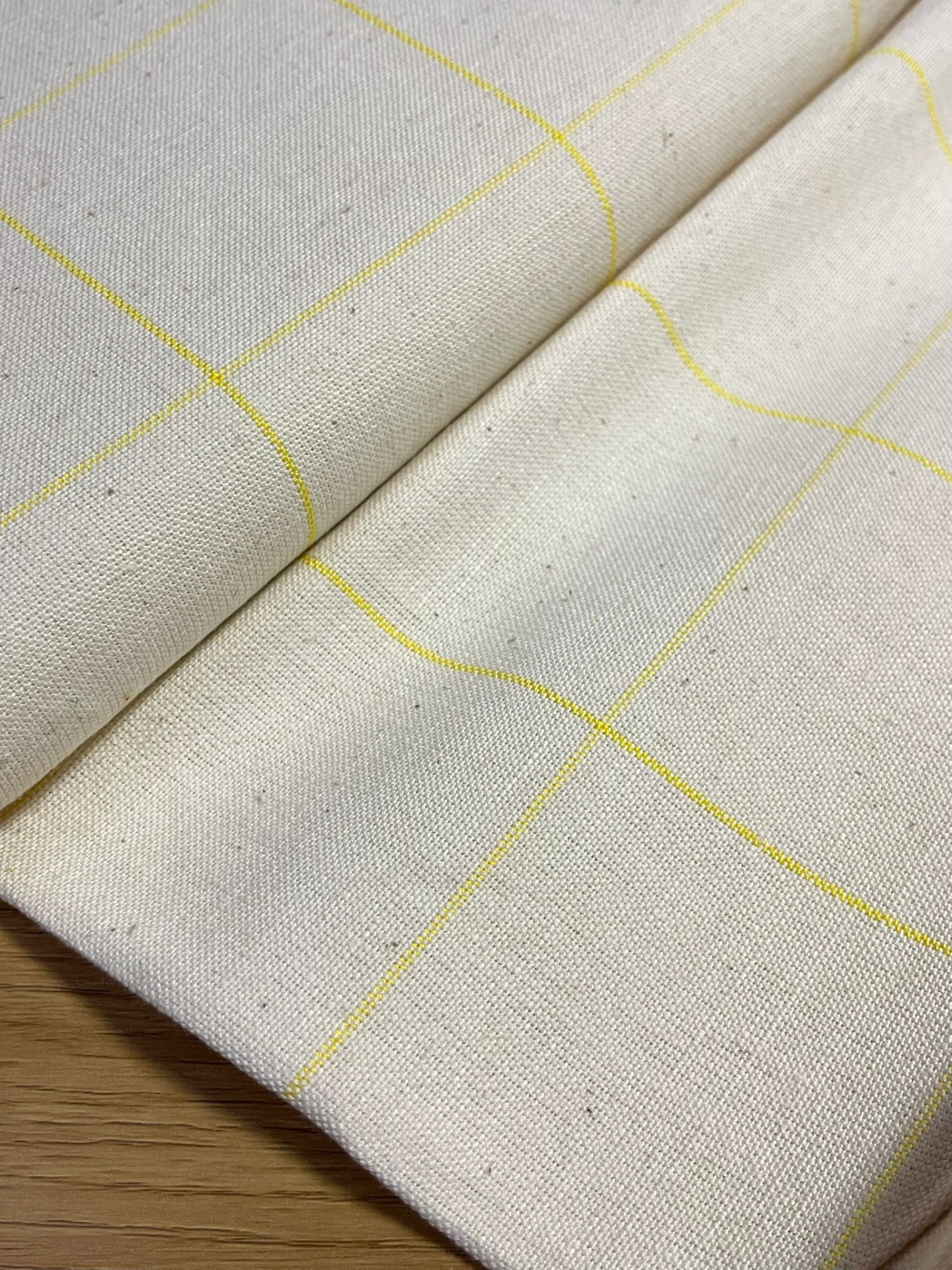 Tecido algodão cru com linhas amarelas em padrão quadriculado, dobrado sobre madeira clara.