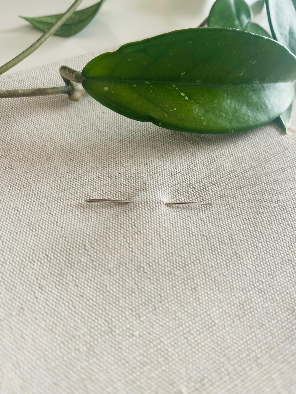 Agulhas de costura em metal sobre tecido branco com folhas verdes ao fundo