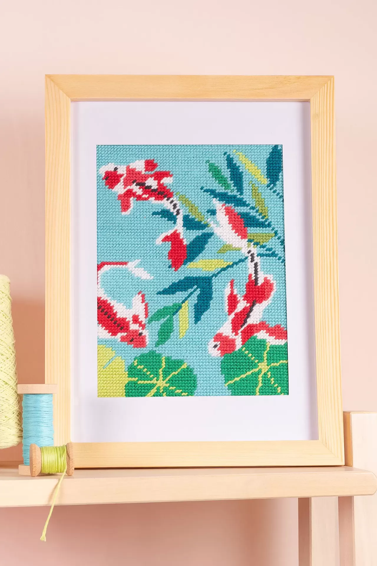 Quadro bordado com peixe koi e folhas em moldura de madeira clara sobre mesa com novelos de linha