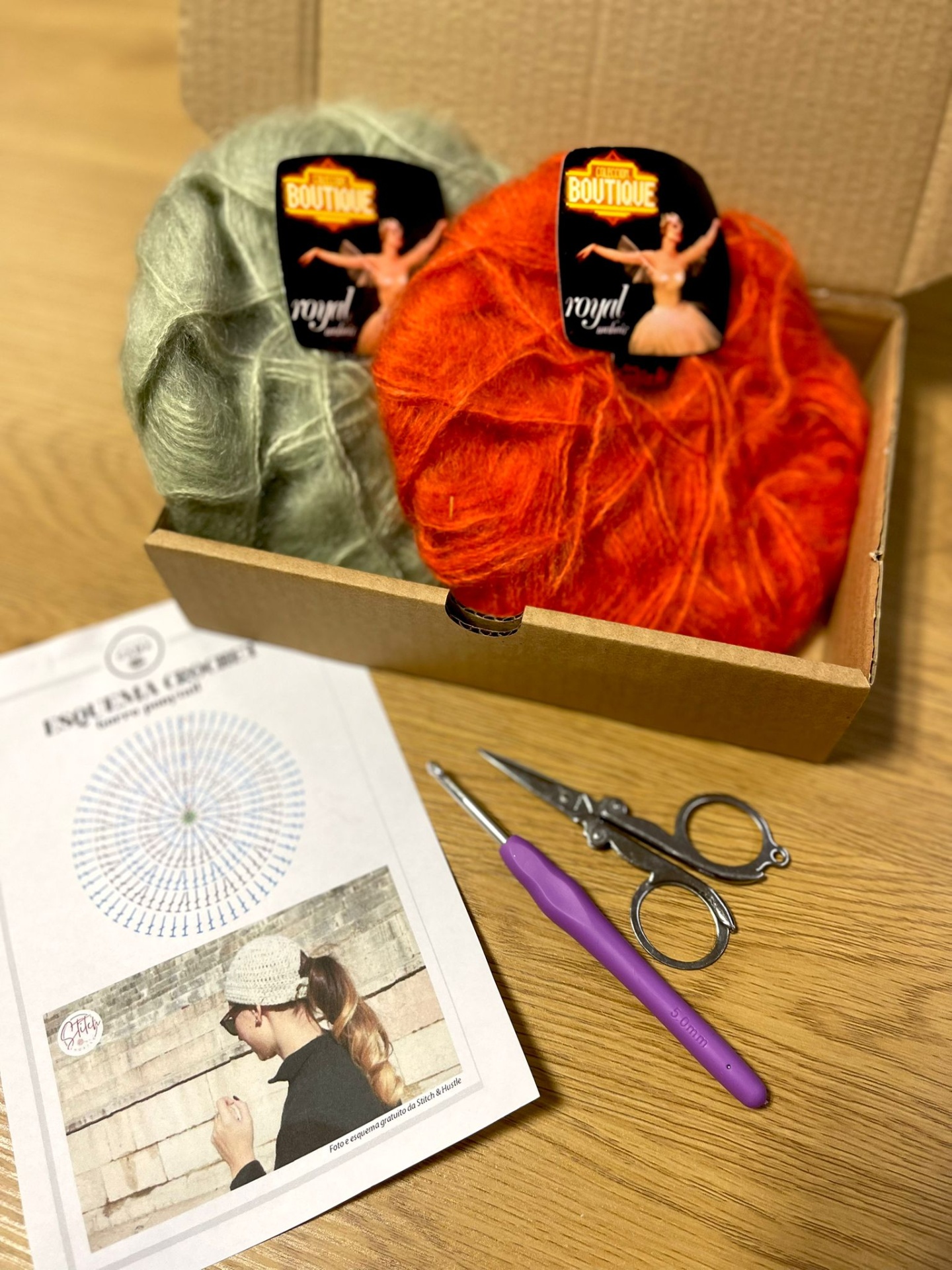 Novelos de lã verde claro e laranja numa caixa com agulha de croché roxa, tesoura e esquema de croché numa folha