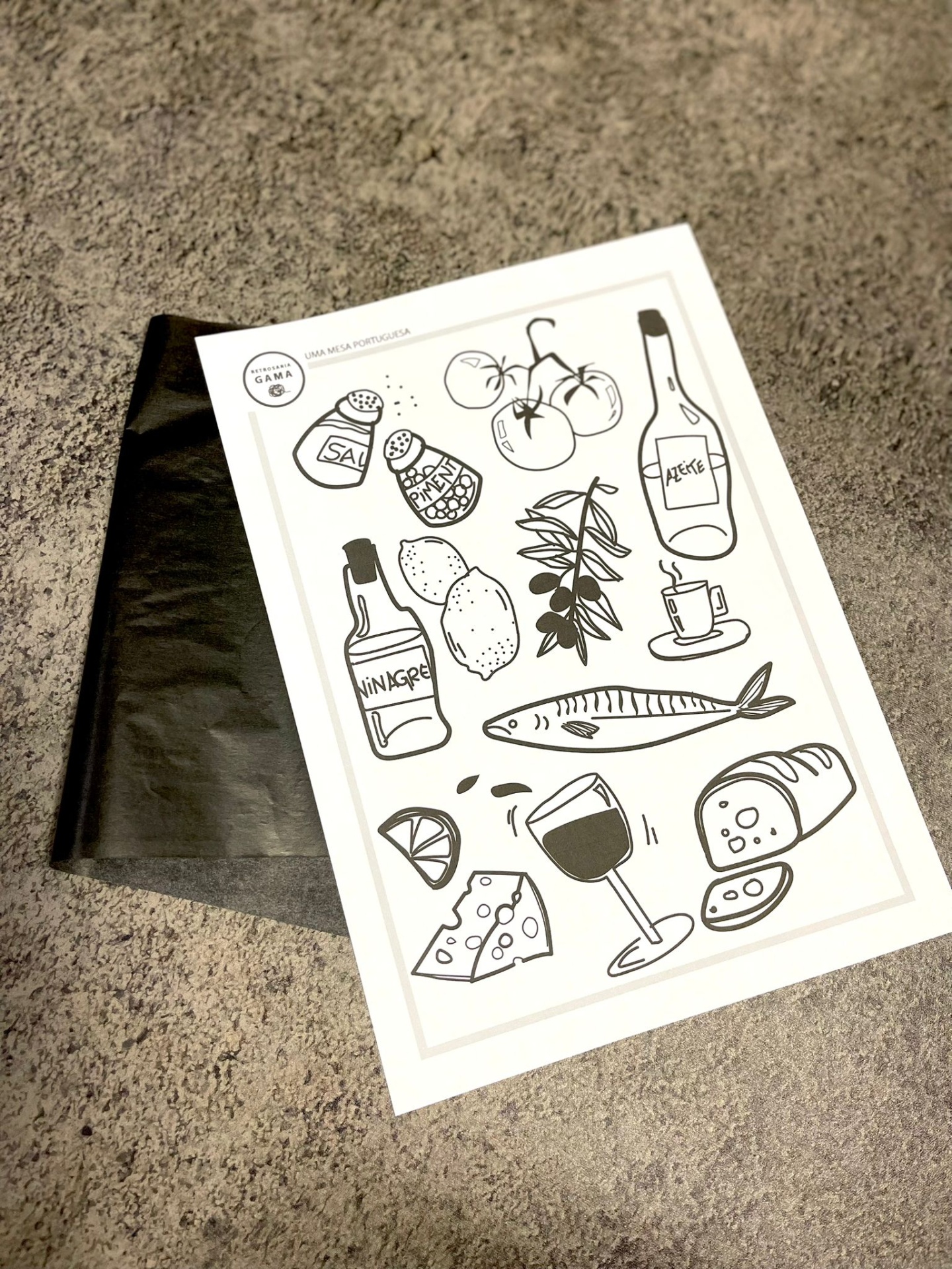 Papel ilustrado com desenhos em preto de alimentos e bebidas sobre superfície cinzenta e saco preto