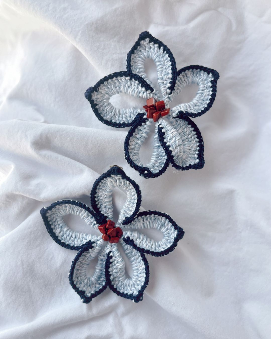 Dois acessórios florais de crochê cinza claro com bordos pretos e pedras vermelhas no centro sobre fundo branco