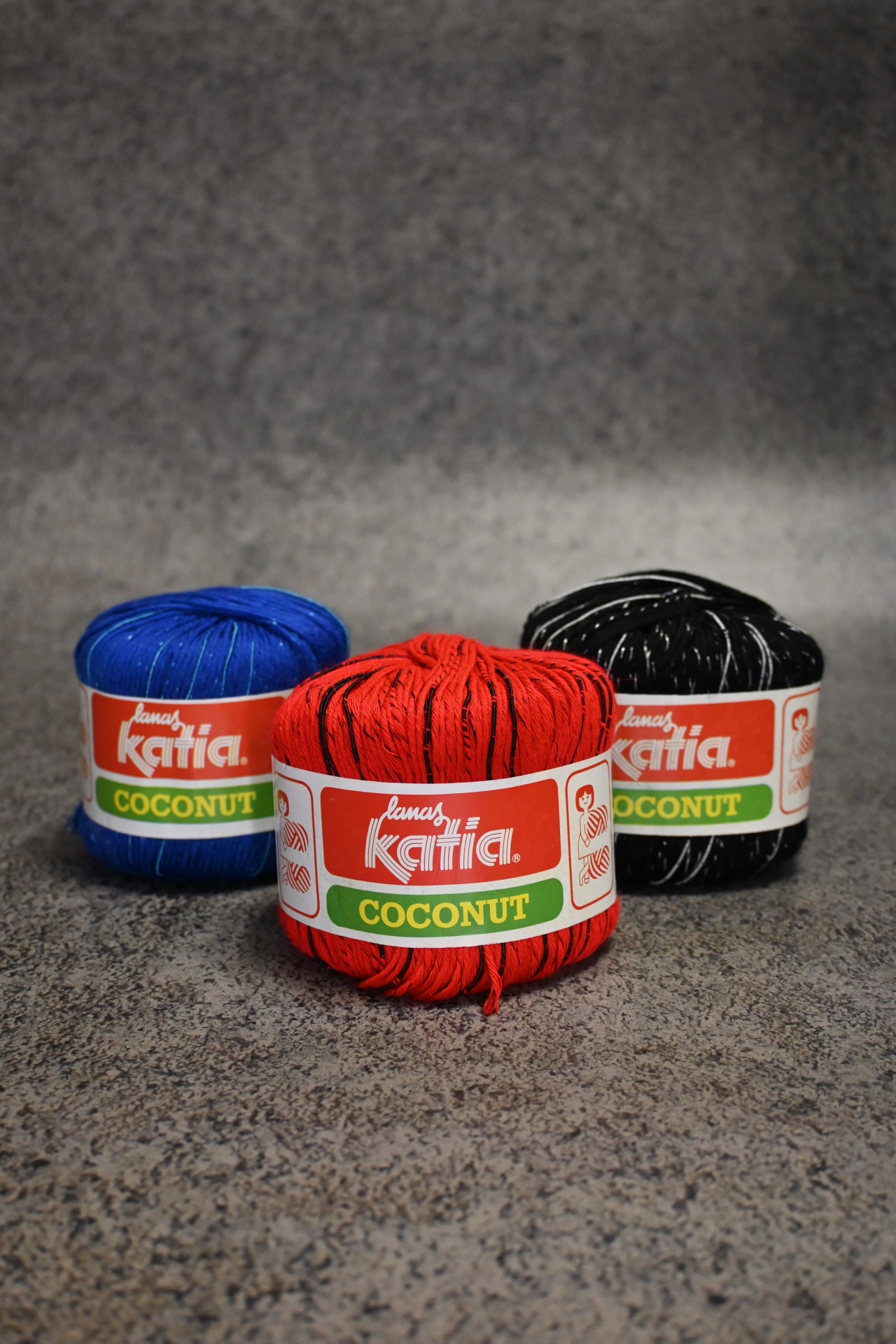 Novelos de lã Katia Coconut em azul, vermelho e preto com rótulos sobre superfície cinzenta.