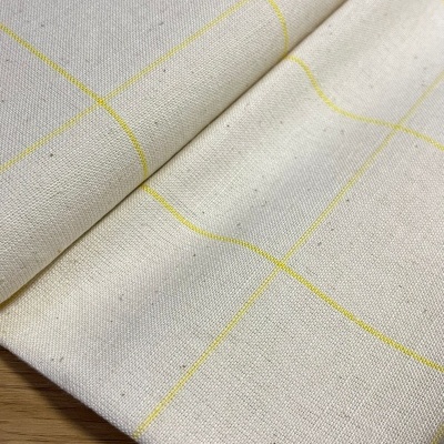 Tecido algodão cru com linhas amarelas em padrão quadriculado, dobrado sobre madeira clara.