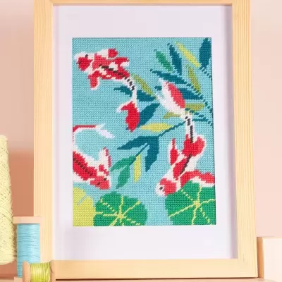 Quadro bordado com peixe koi e folhas em moldura de madeira clara sobre mesa com novelos de linha