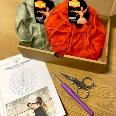 Novelos de lã verde claro e laranja numa caixa com agulha de croché roxa, tesoura e esquema de croché numa folha