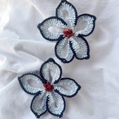 Dois acessórios florais de crochê cinza claro com bordos pretos e pedras vermelhas no centro sobre fundo branco