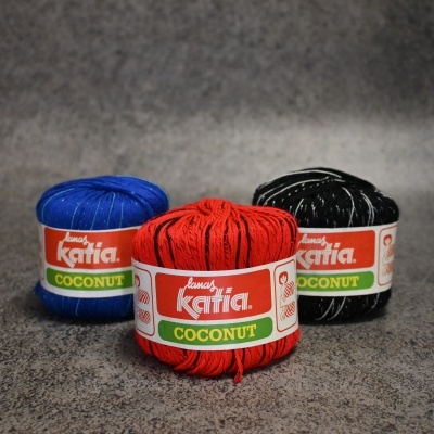 Novelos de lã Katia Coconut em azul, vermelho e preto com rótulos sobre superfície cinzenta.