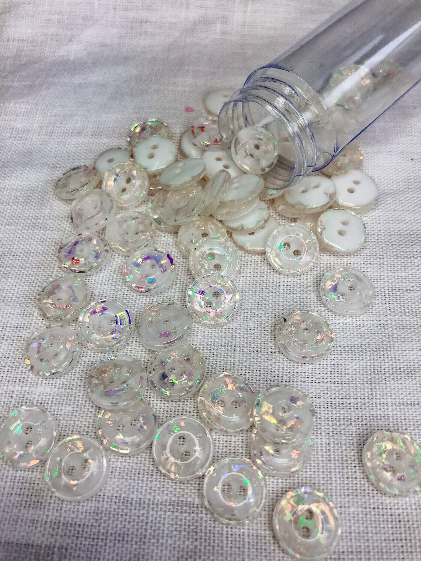 Botões transparentes iridescentes e brancos sobre tecido branco ao lado de tubo plástico.