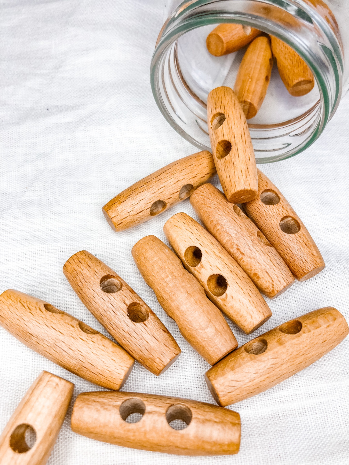 Botões de madeira cor de mel, cilíndricos, com furos, sobre tecido branco e frasco de vidro