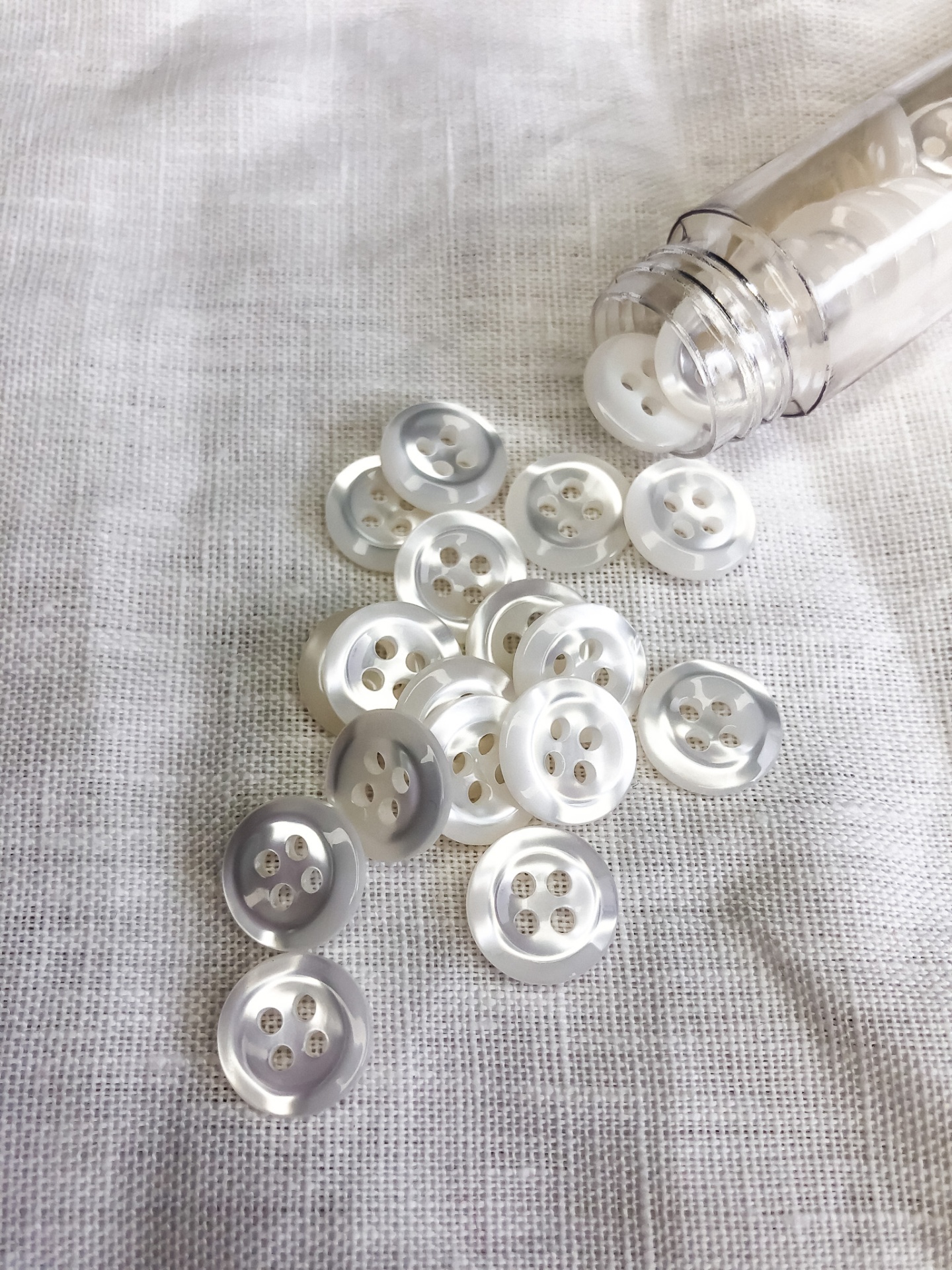 Botões transparentes com quatro furos espalhados em pano branco junto a um frasco transparente aberto