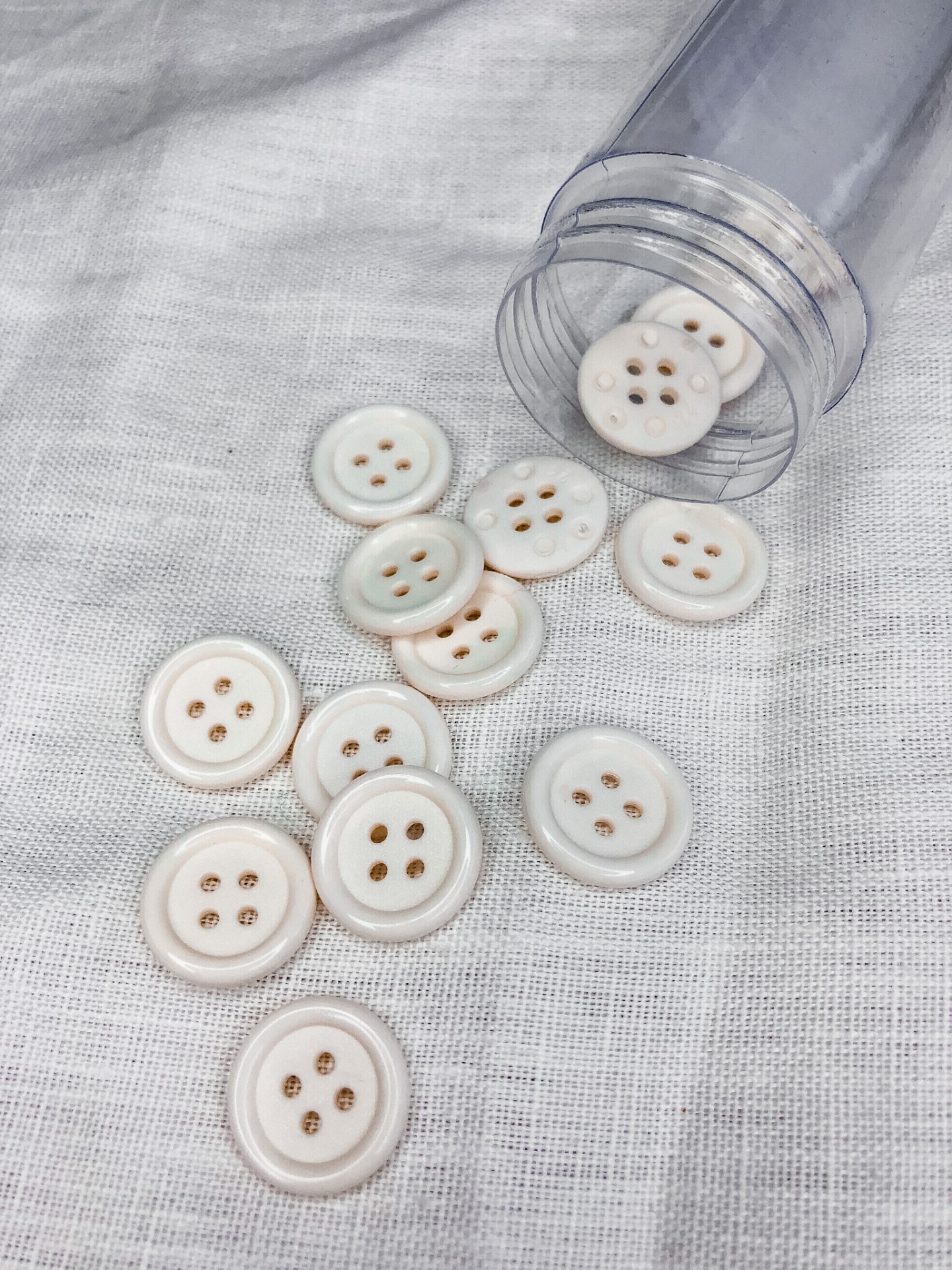 Botões brancos redondos com quatro furos sobre tecido branco e recipiente plástico transparente.