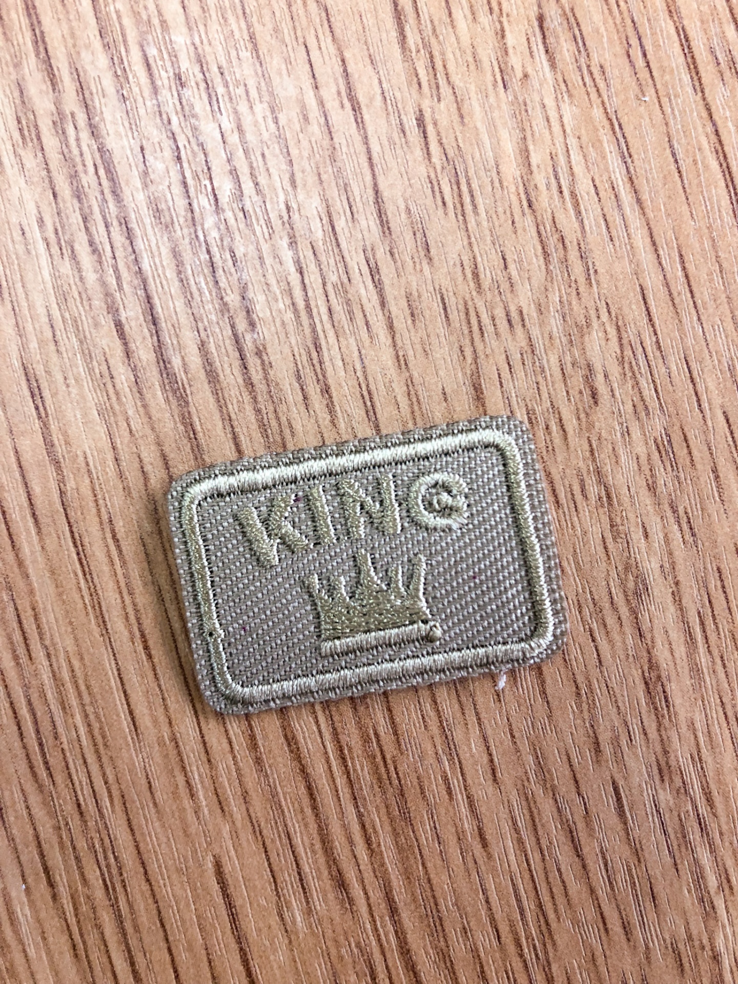 Etiqueta têxtil bege com 'KING' e coroa sobre madeira clara