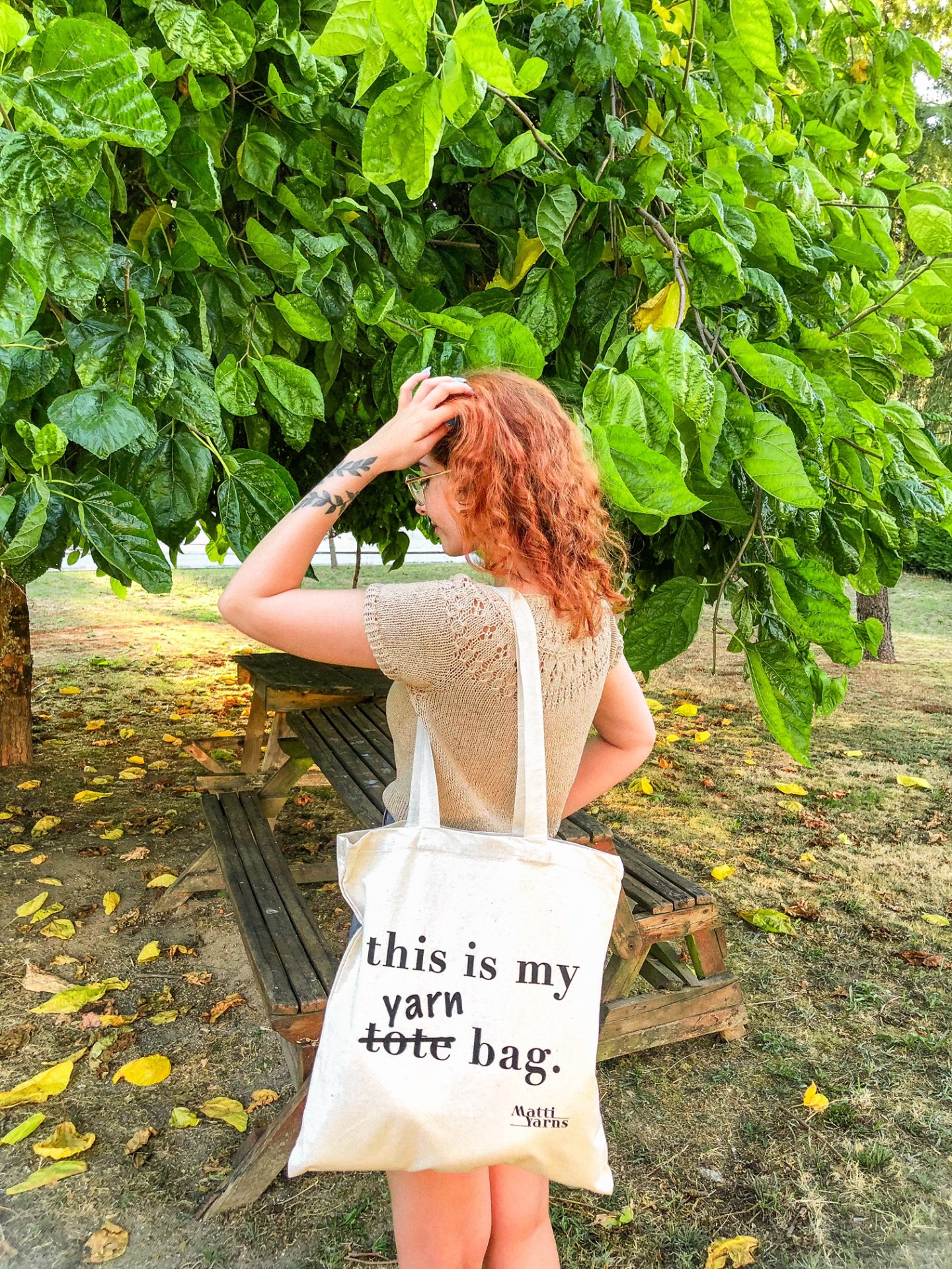 Saco de pano bege com texto preto usado por mulher ruiva sentada em banco de madeira numa área ao ar livre com muitas folhas verdes.