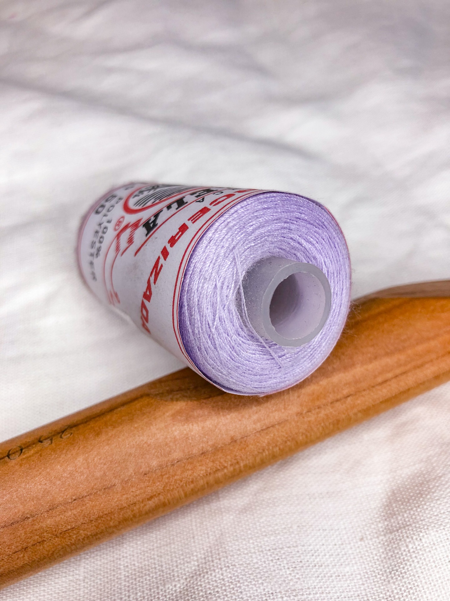 Carrete de linha lilás sobre tecido branco e cabo de madeira.