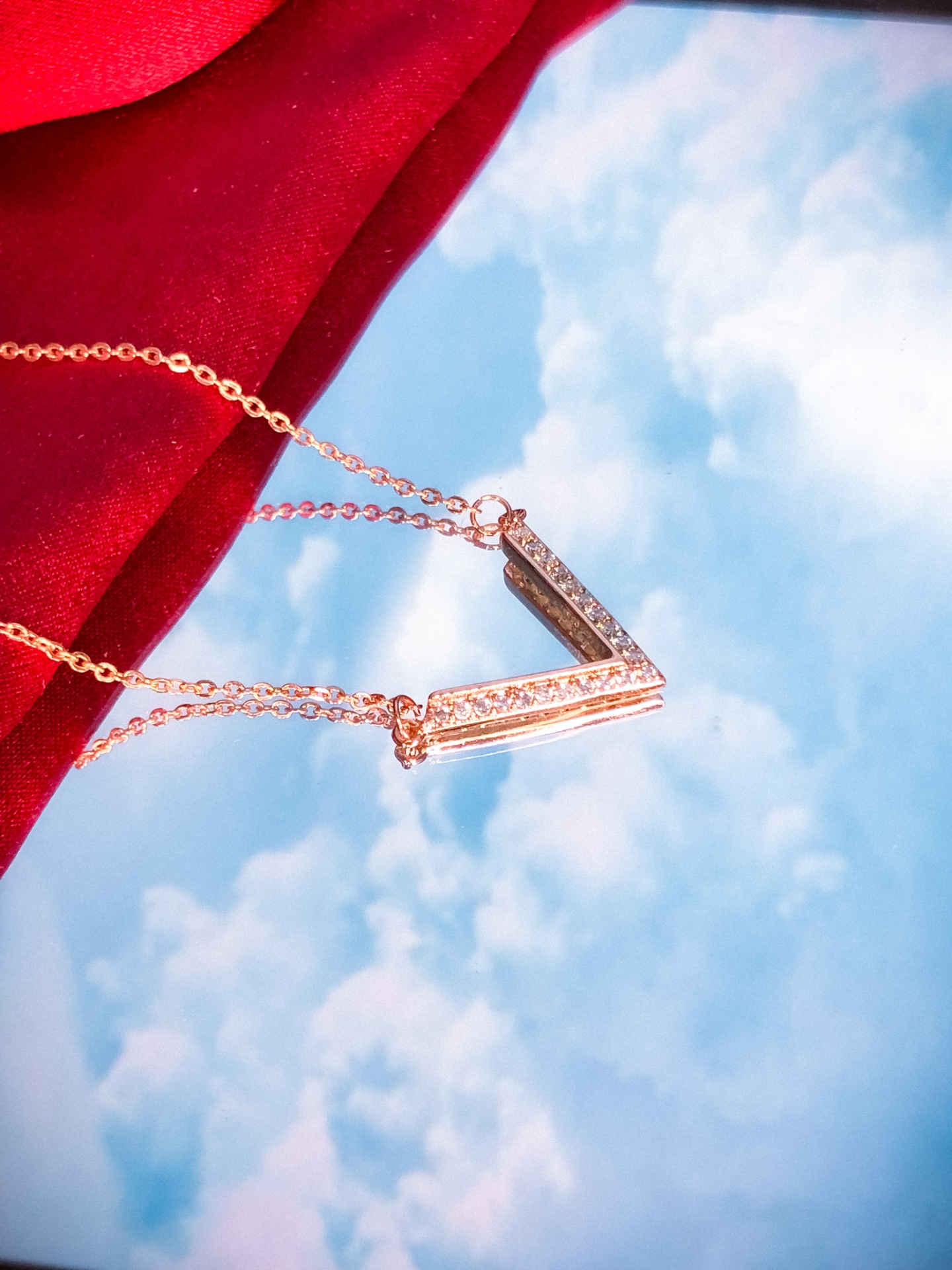 Colar dourado com pendente em V e pedras brilhantes sobre fundo vermelho e céu azul refletido