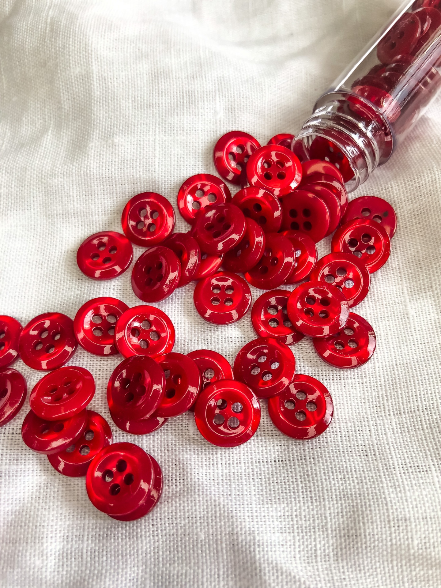 Botões vermelhos com quatro furos sobre tecido branco, saindo de recipiente transparente.