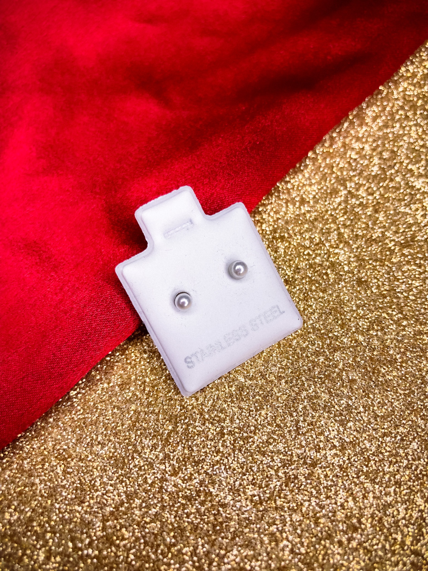 Brincos de aço inoxidável em suporte branco sobre fundo dourado e vermelho.