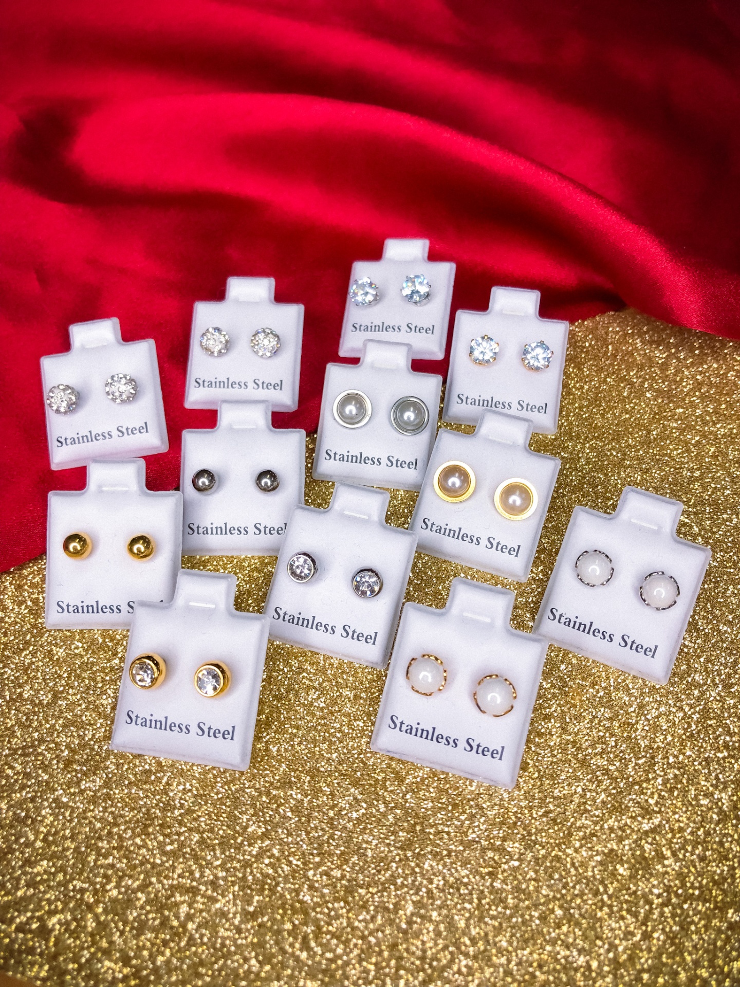 Conjunto de brincos em aço inoxidável com pedras, em suportes brancos, sobre fundo dourado e vermelho