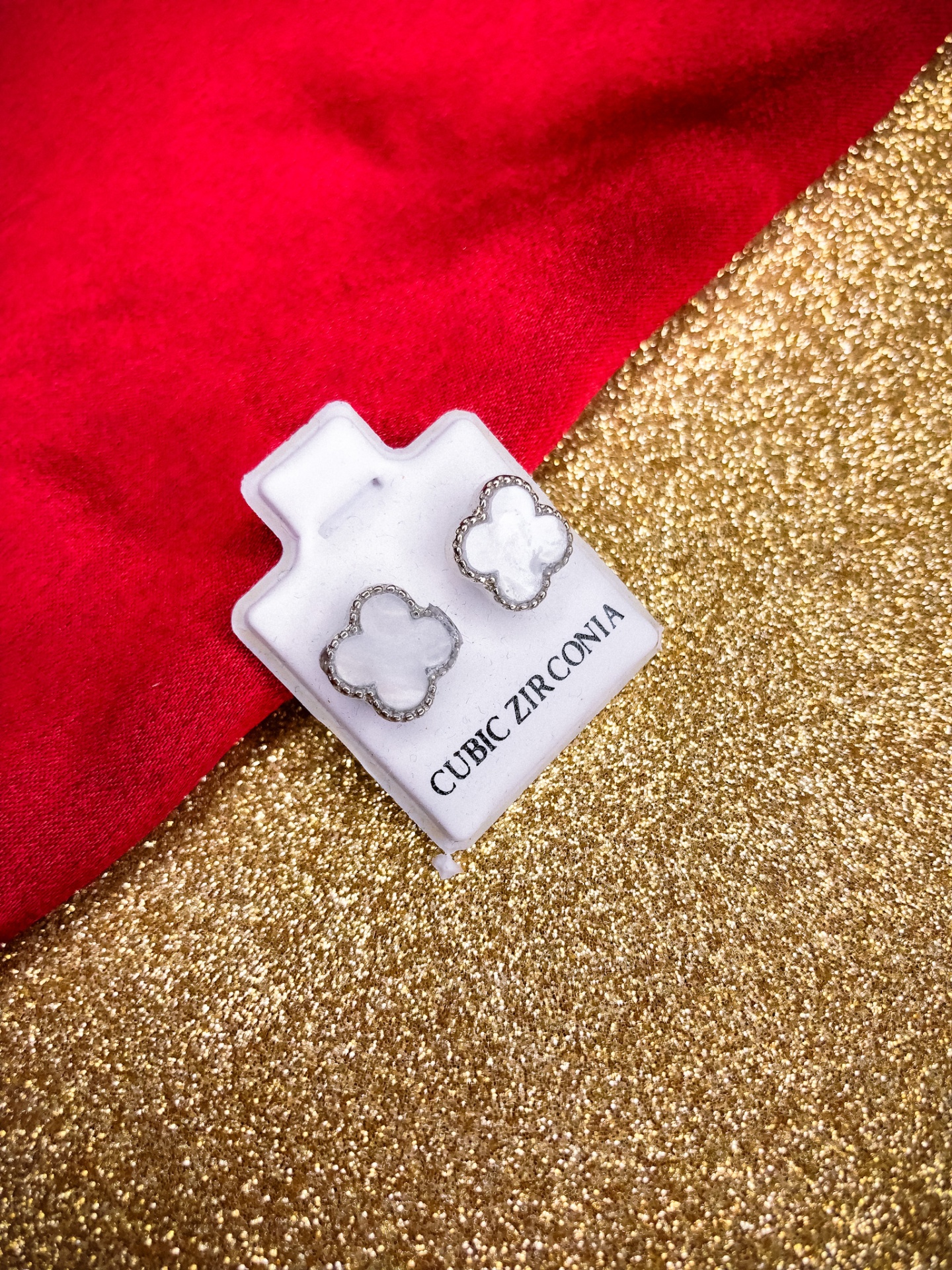 Brincos florais de madrepérola com contorno prateado num suporte branco com texto, fundo dourado e tecido vermelho