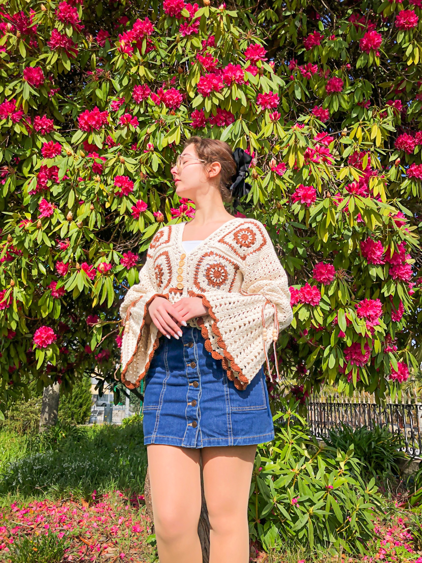 Mulher com blusa de malha bege e saia de ganga azul em frente a arbusto com flores cor de rosa