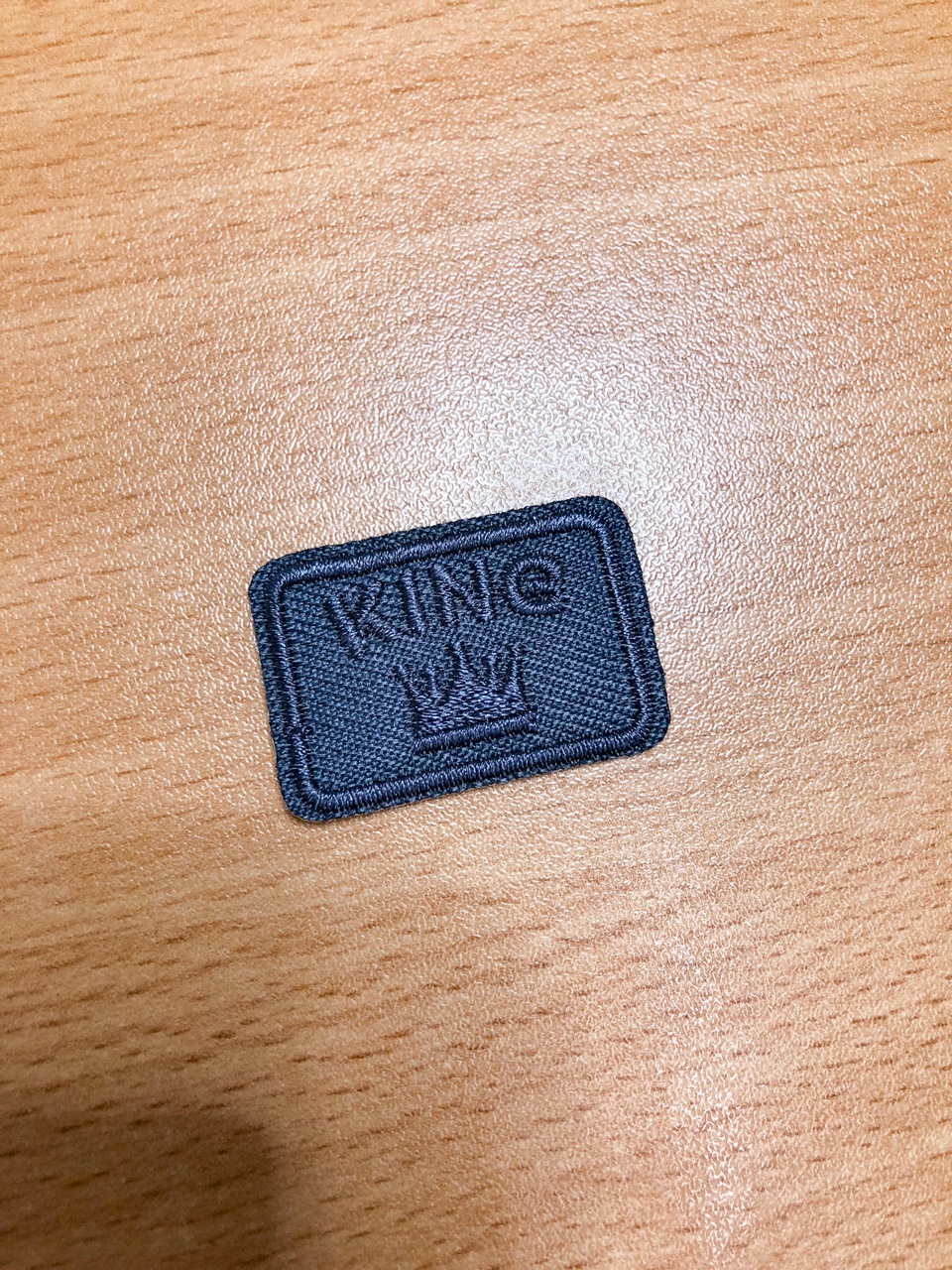 Etiqueta de tecido preta com texto KING e coroa sobre madeira clara