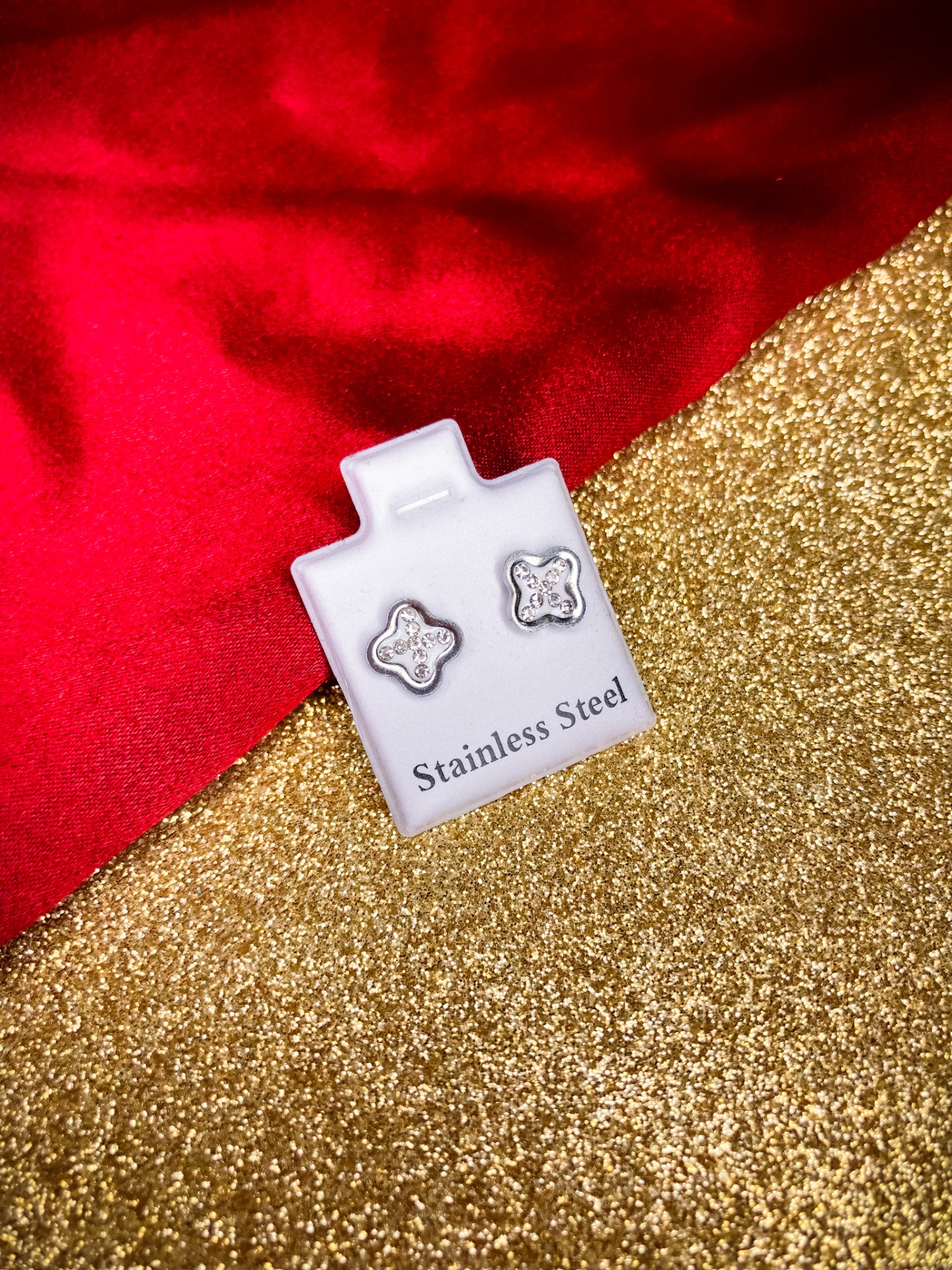 Brincos de aço inoxidável em forma de flor com cristais num suporte branco com texto, sobre fundo dourado e vermelho