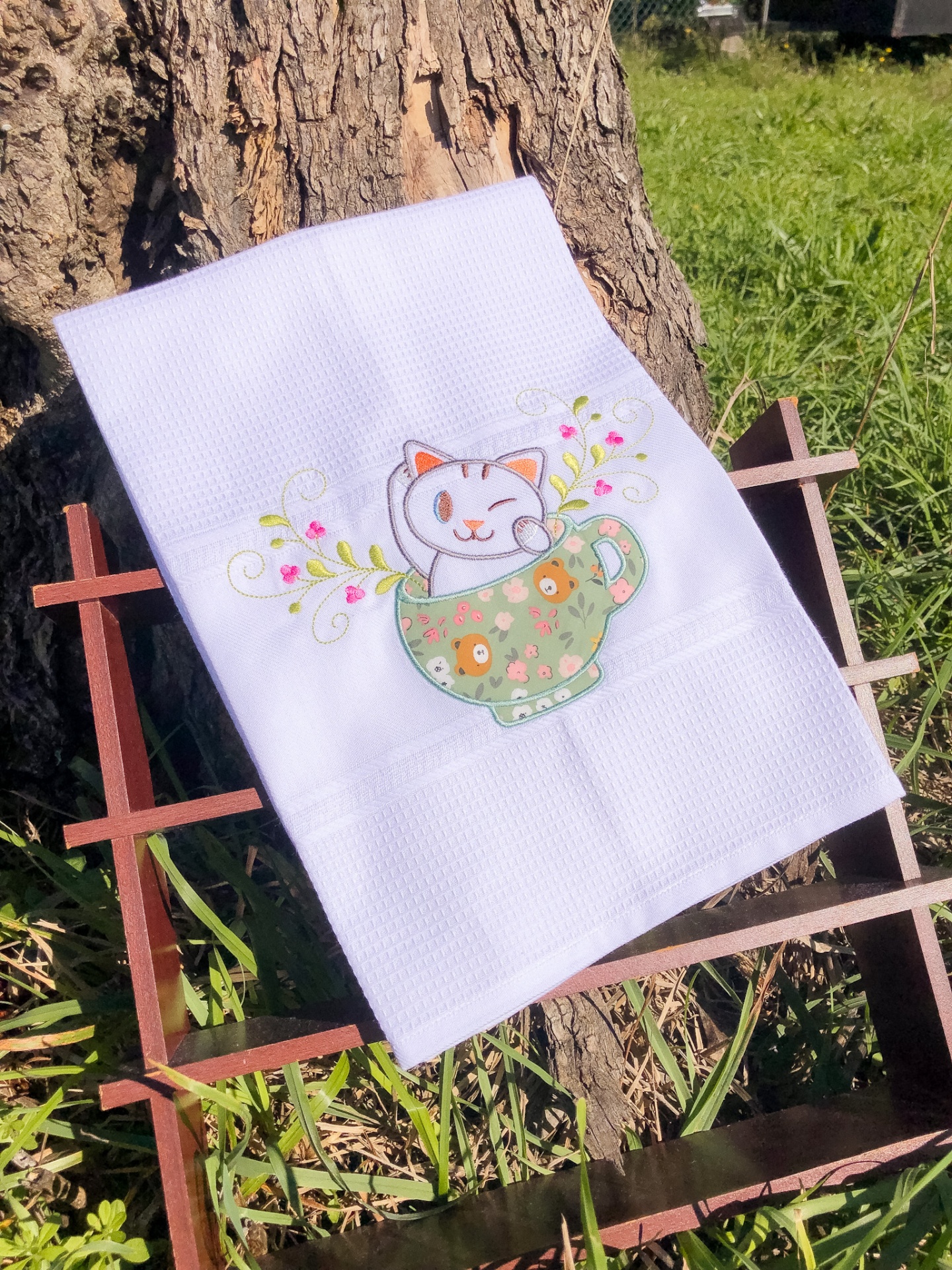 Toalha branca com bordado de gato numa chávena verde suportada por estrutura de madeira ao ar livre