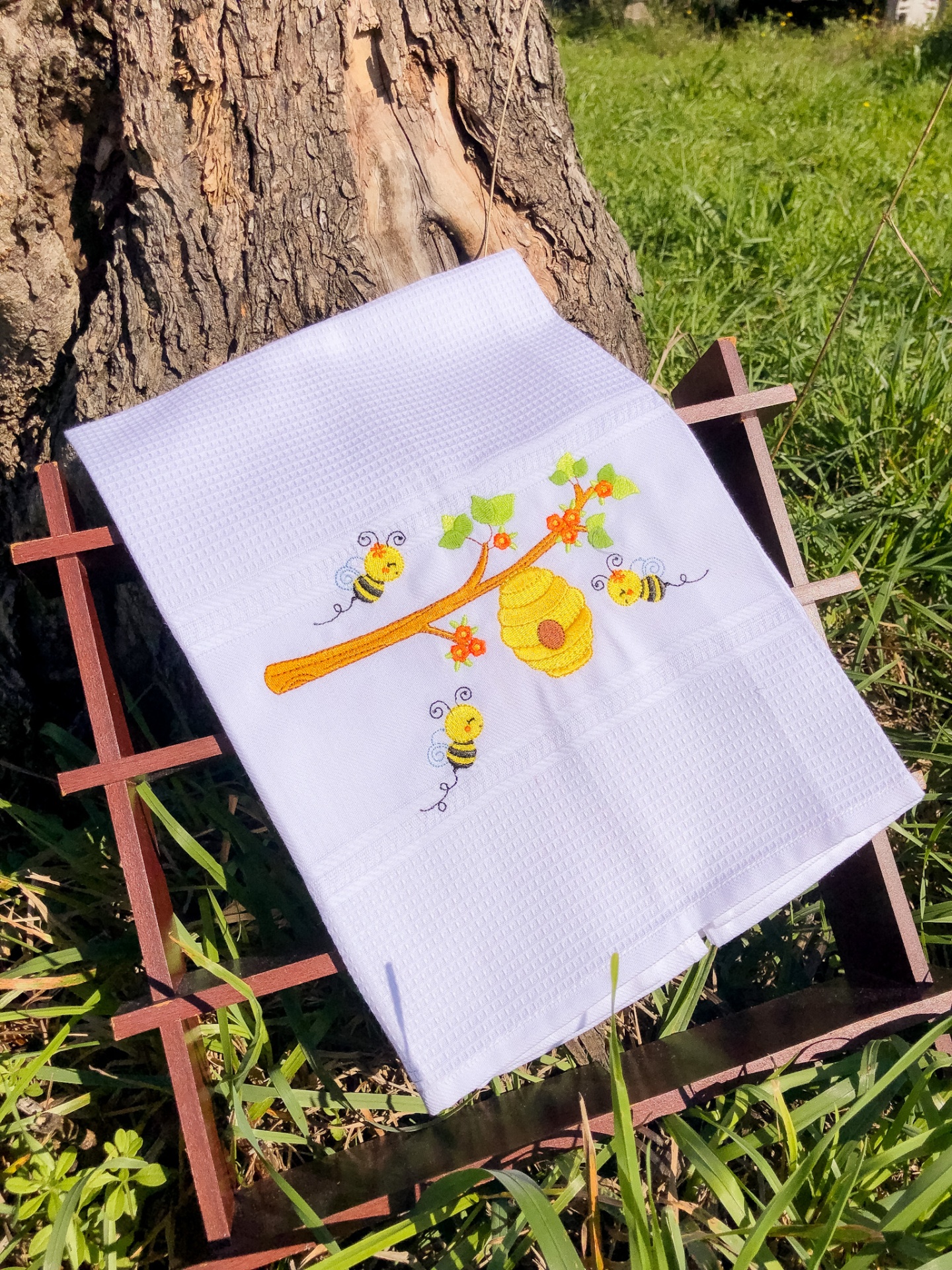 Toalha branca bordada com favo de mel e abelhas sobre estrutura de madeira na relva
