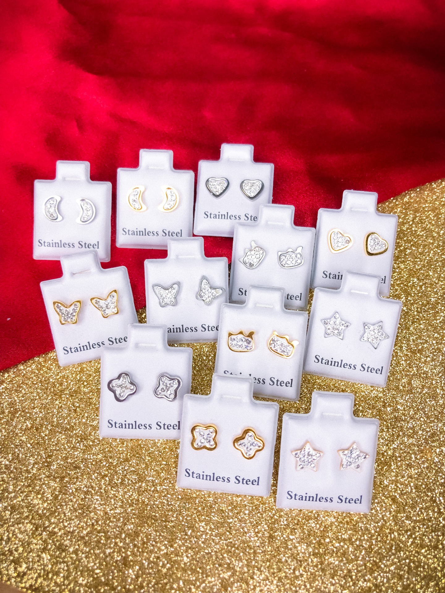 Conjunto de brincos de aço inoxidável em diversas formas e cores, em cartões brancos sobre tecido vermelho e dourado.