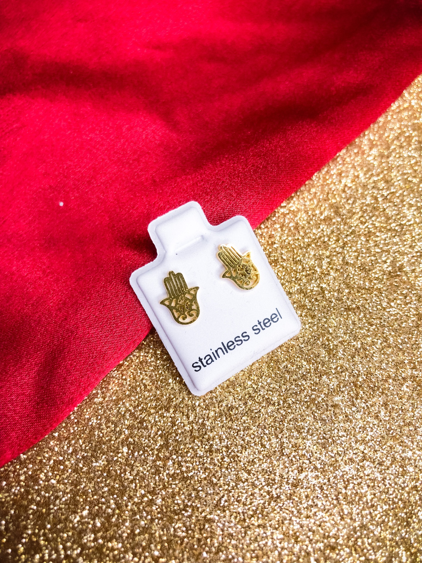 Brincos dourados em forma de mão da Fátima sobre suporte branco com fundo vermelho e dourado