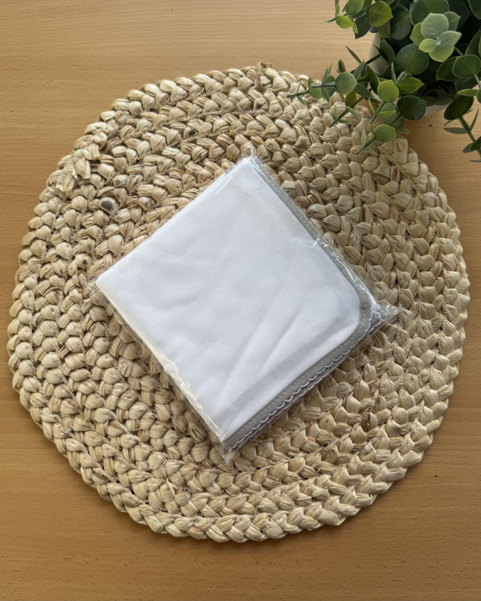 Pano branco embalado em plástico sobre suporte redondo tecido natural com planta verde ao lado