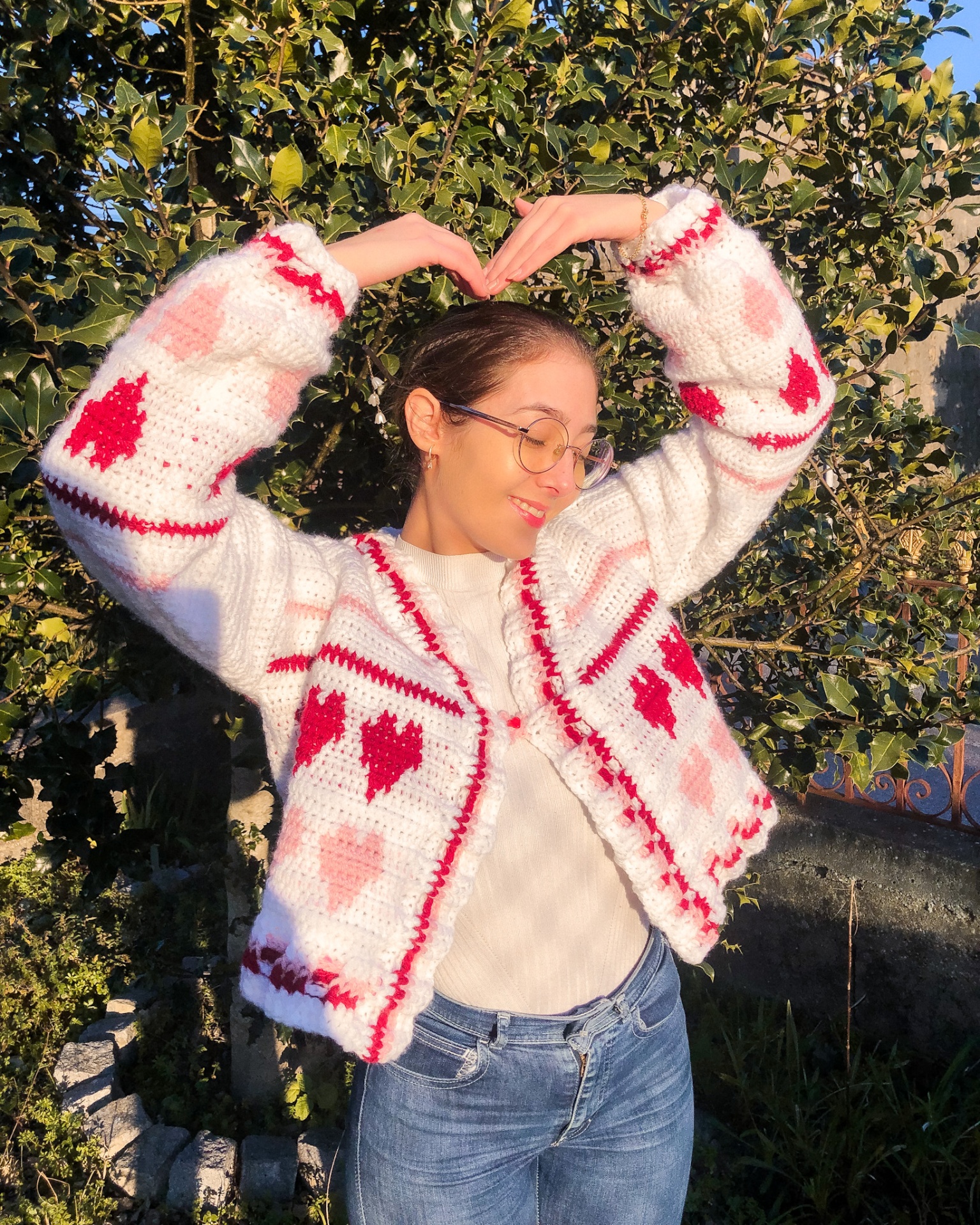 Mulher com casaco de malha branco com corações vermelhos e rosa, óculos, blusa branca e jeans, ao ar livre
