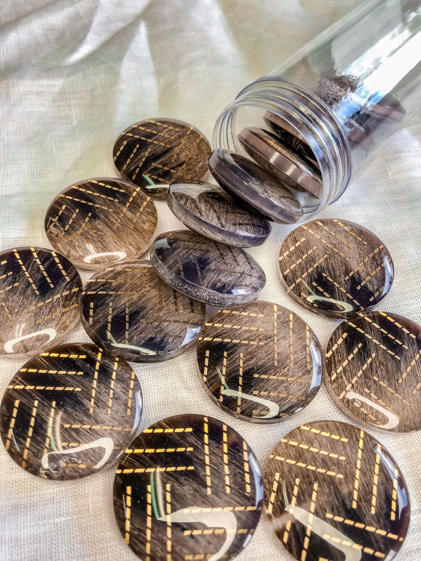 Botões redondos com padrão preto e dourado derramados de um recipiente plástico sobre tecido claro