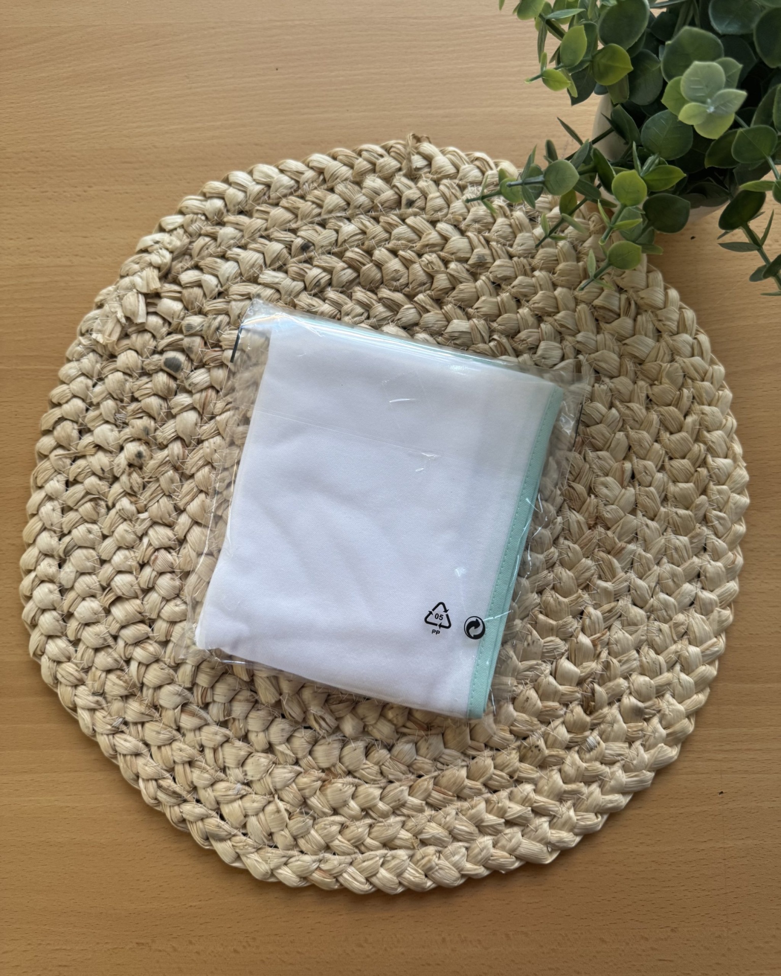 pano branco com borda azul claro embalado em plástico sobre descanso redondo de palha e mesa de madeira clara