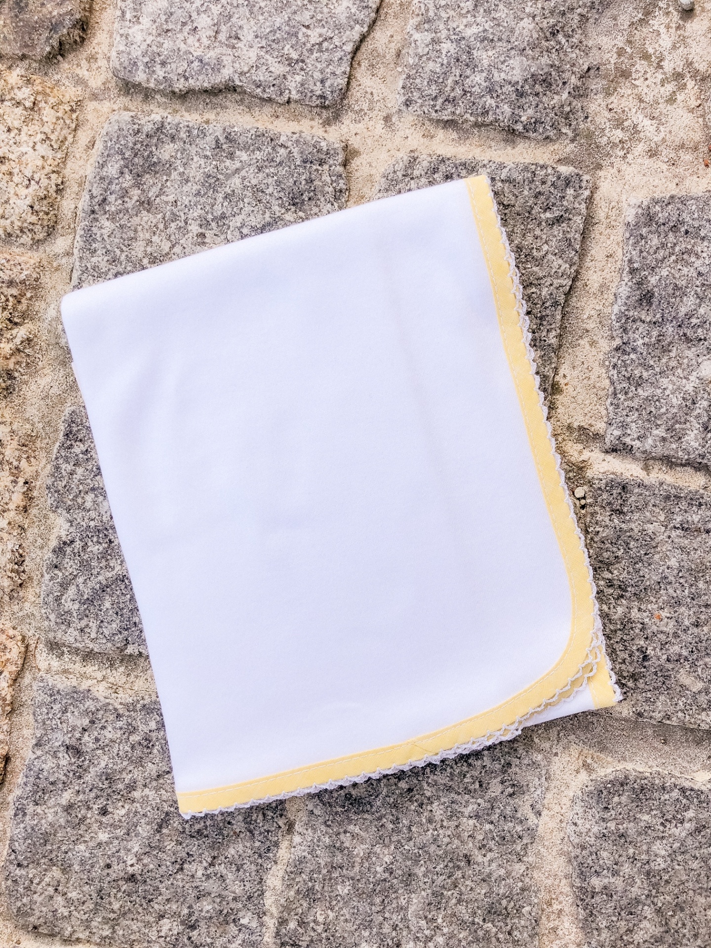 Lenço branco com borda amarela e renda sobre calçada de pedra.