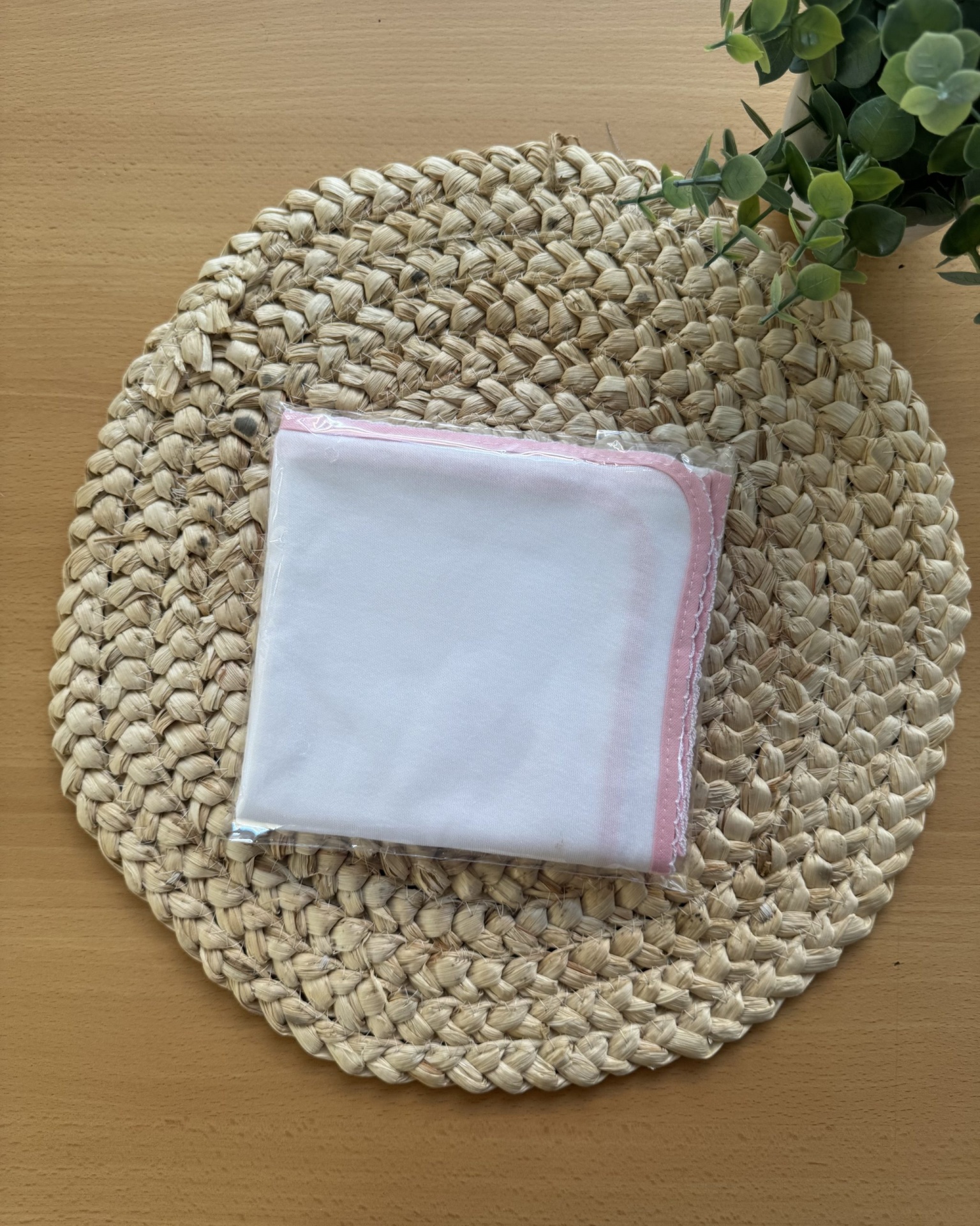 Toalha branca com borda rosa clara dentro de embalagem plástica sobre placemat trançado redondo