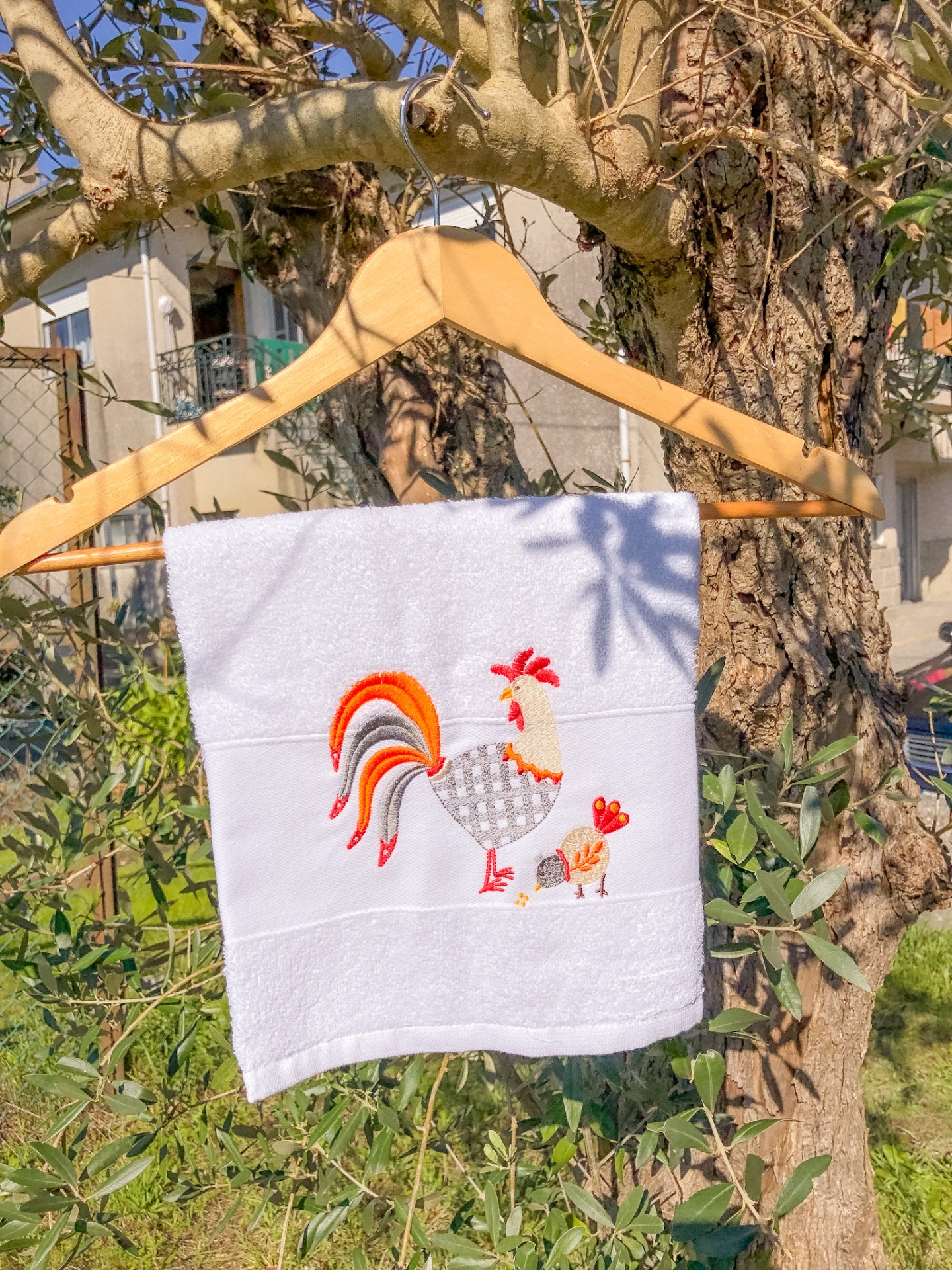 Toalha branca bordada com galo e pintainho pendurada numa cabide de madeira em árvore