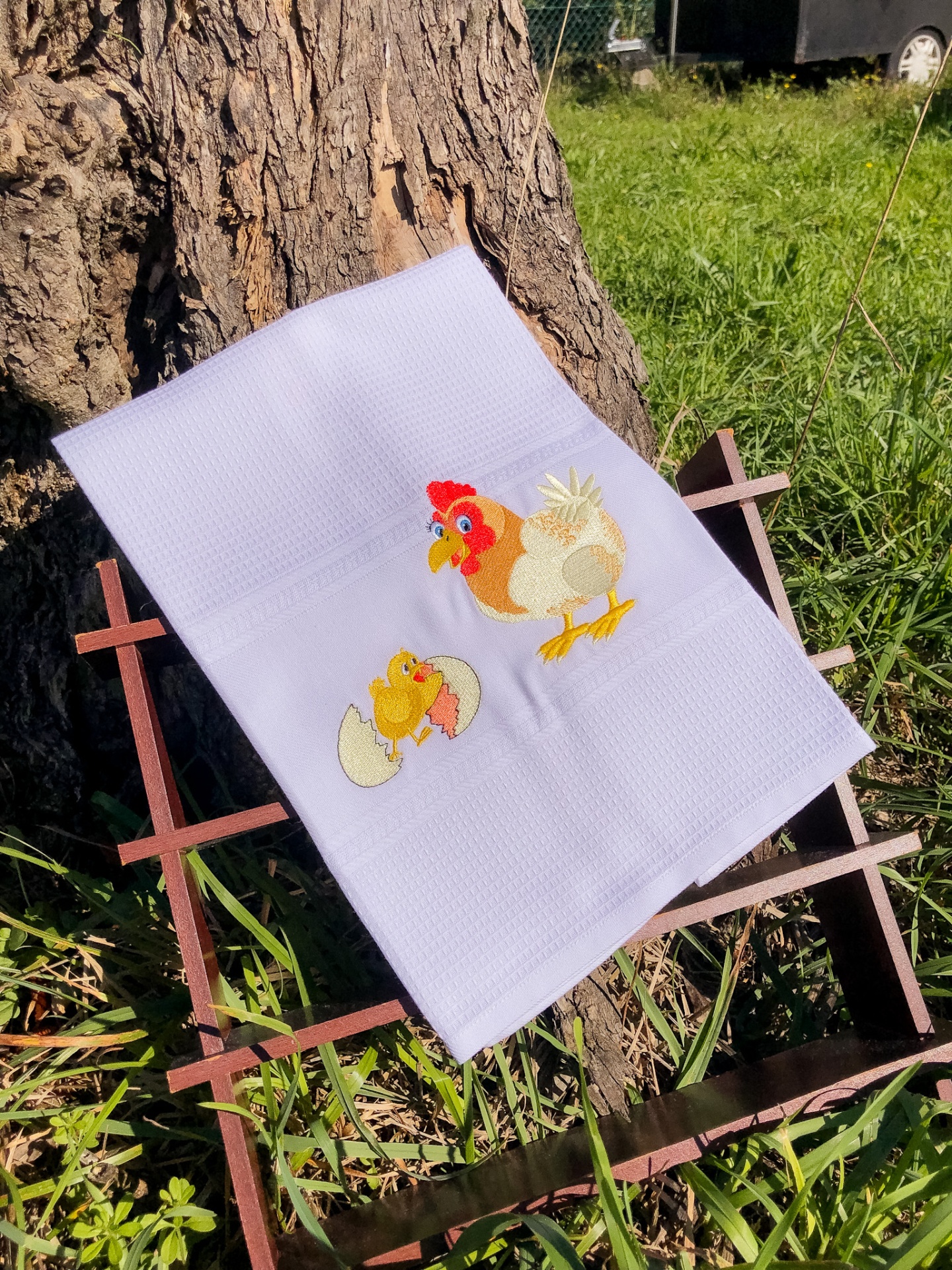 Toalha branca com bordado de galinha e pintainho sobre estrutura de madeira junto a um tronco de árvore.
