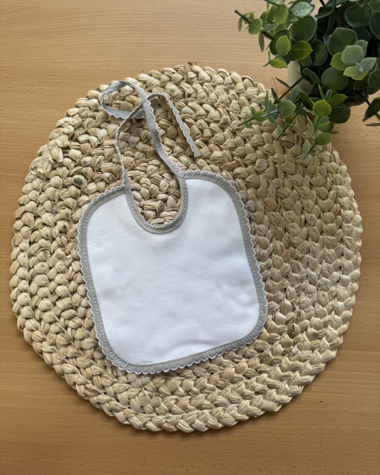 Babete branco com borda cinza sobre suporte trançado bege e superfície de madeira com planta
