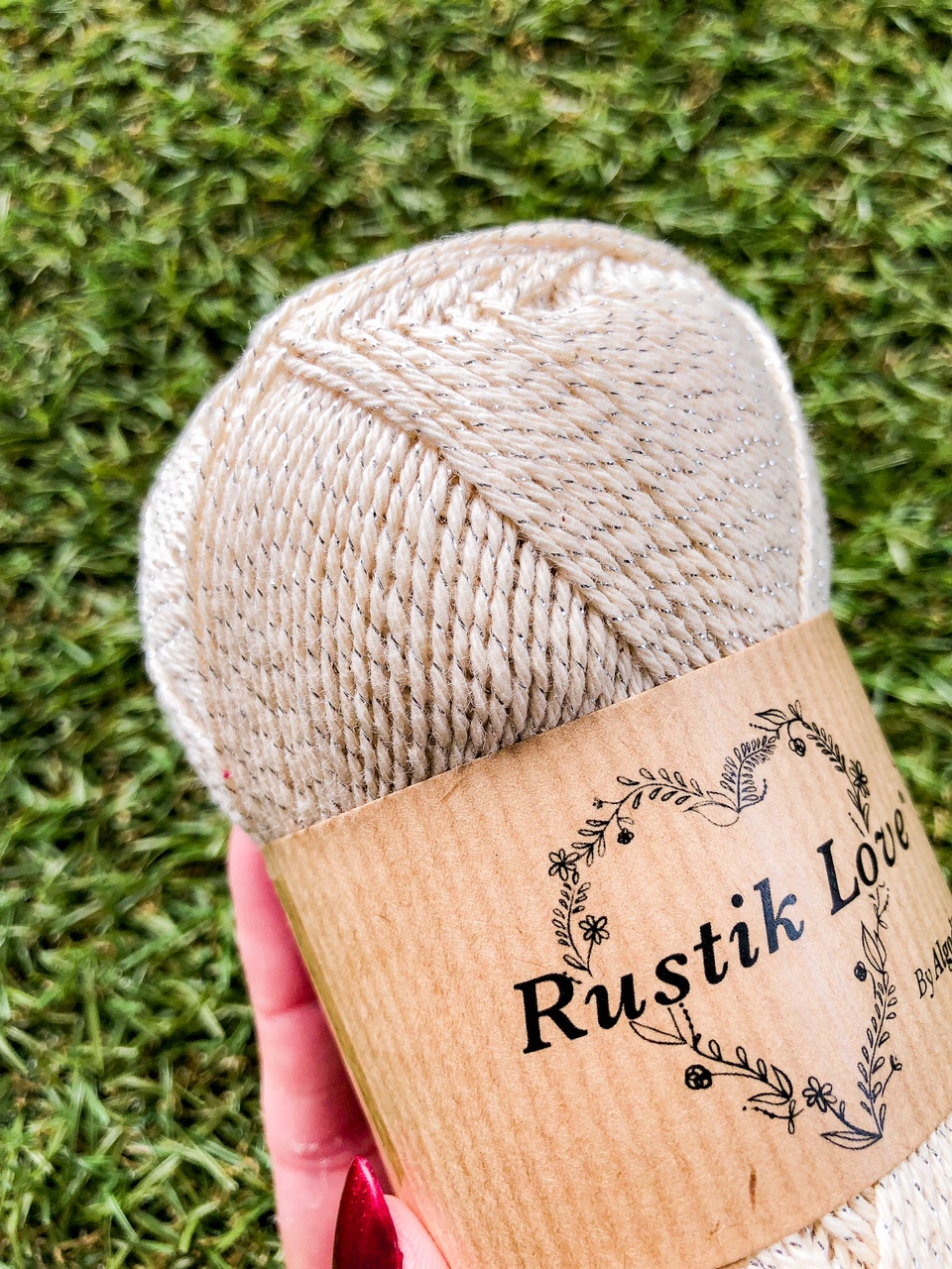 Novelo de fio bege com rótulo Rustik Love segurado em mão com unhas vermelhas sobre relva