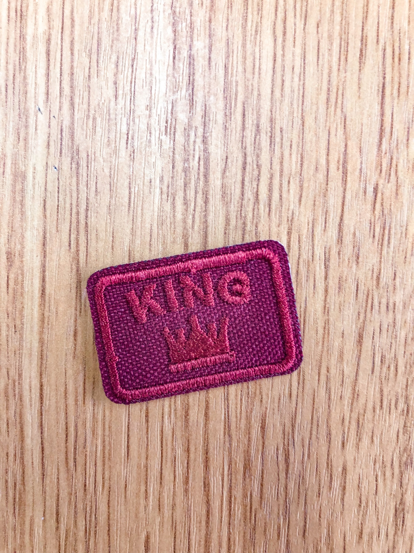 Emblema bordado sentido KING com coroa em tecido vinho numa superfície de madeira clara
