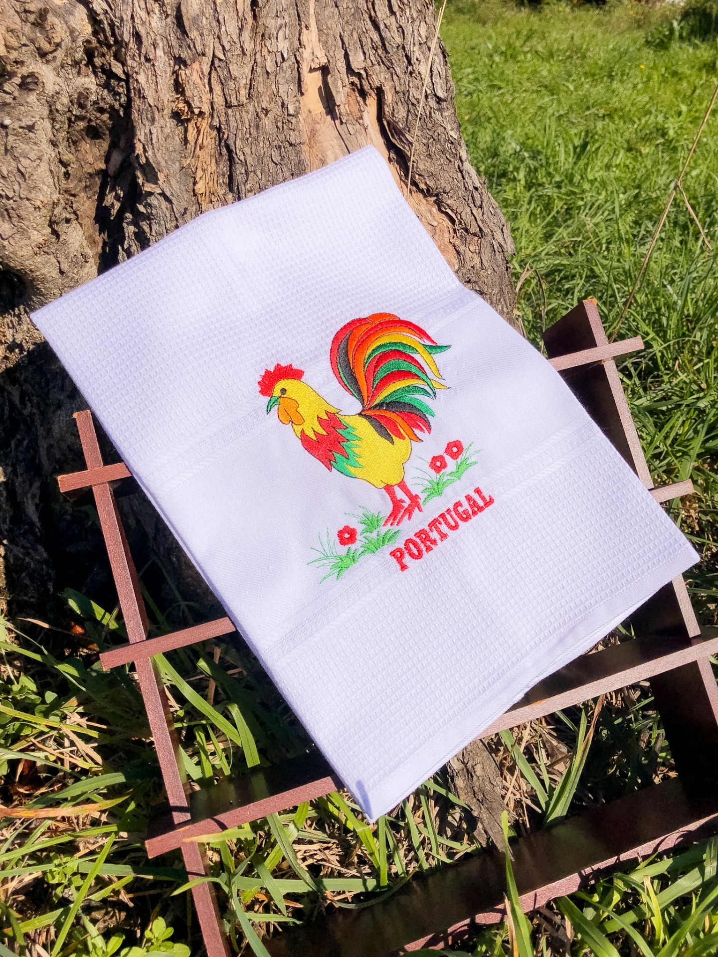 Toalha branca com galo colorido bordado e a palavra PORTUGAL junto a tronco de árvore