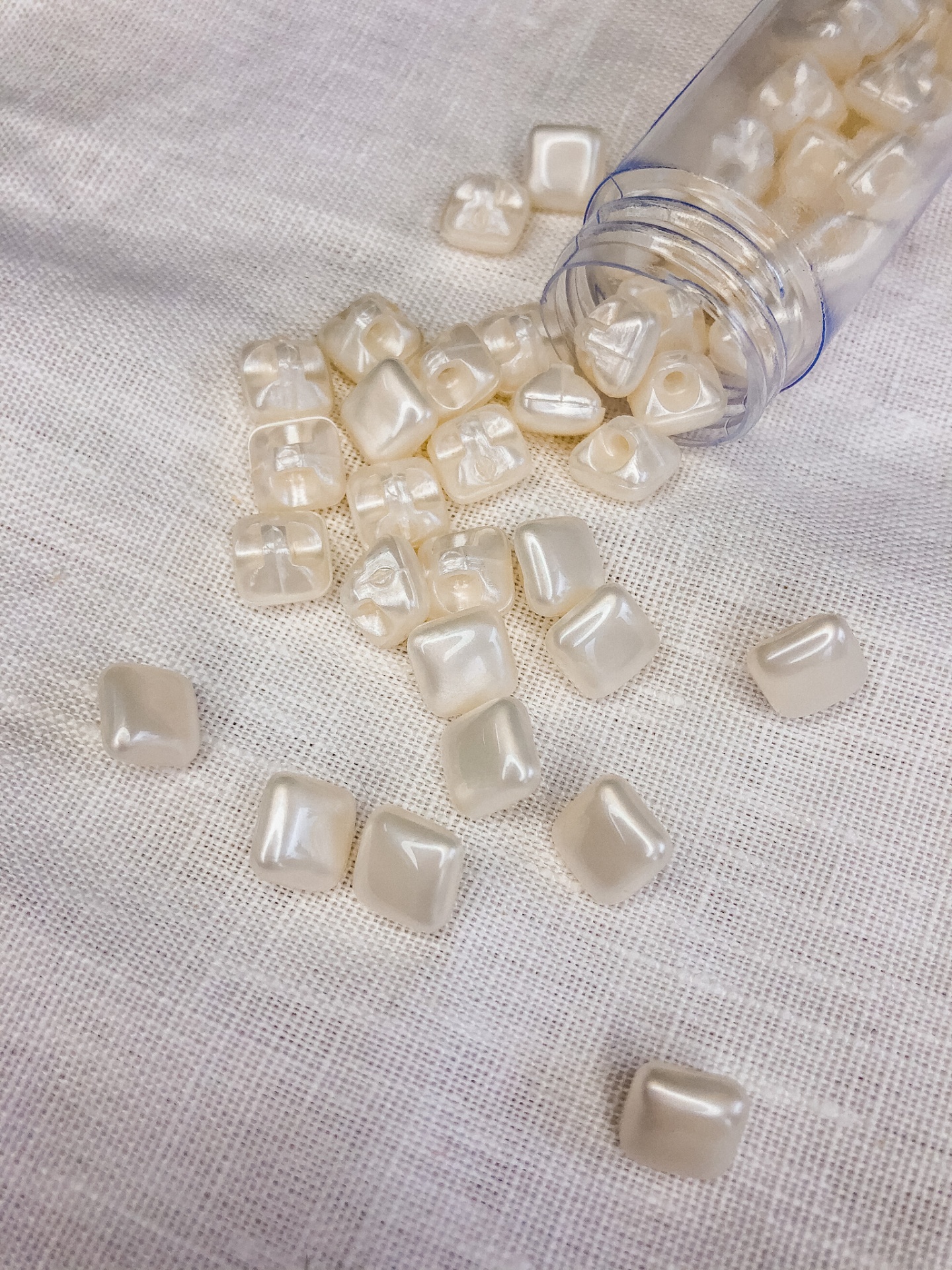 Contas quadradas peroladas brancas sobre tecido branco com garrafa transparente tombada