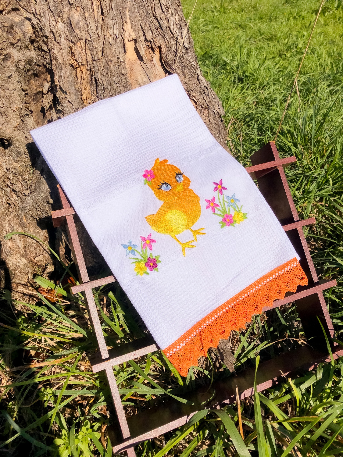 Toalha branca com bordado de pintainho amarelo e flores, com borda de croché laranja, sobre suporte de madeira ao ar livre