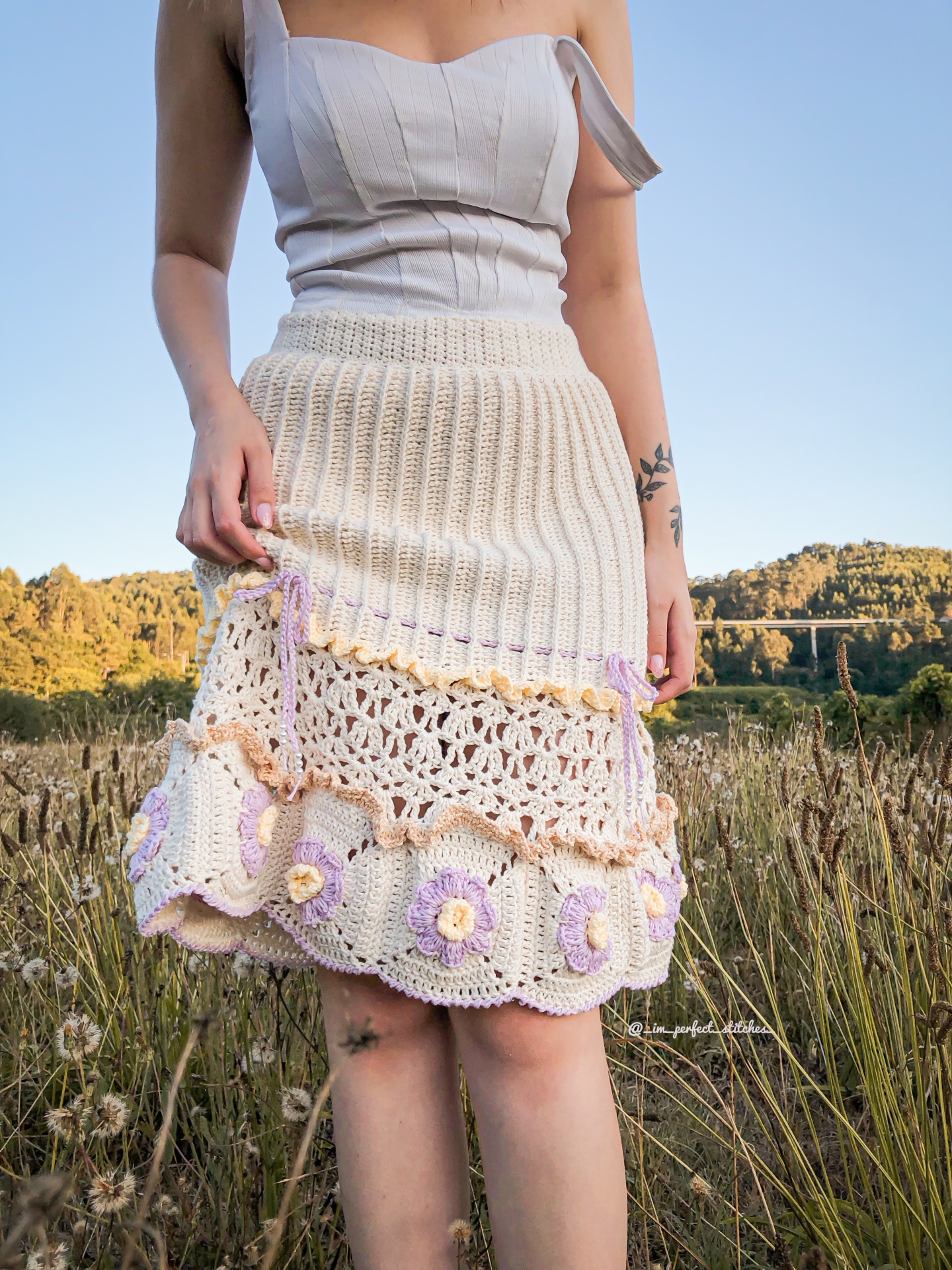 Saia de malha branca com detalhes em crochê floral lilás e amarelo, usada numa paisagem natural