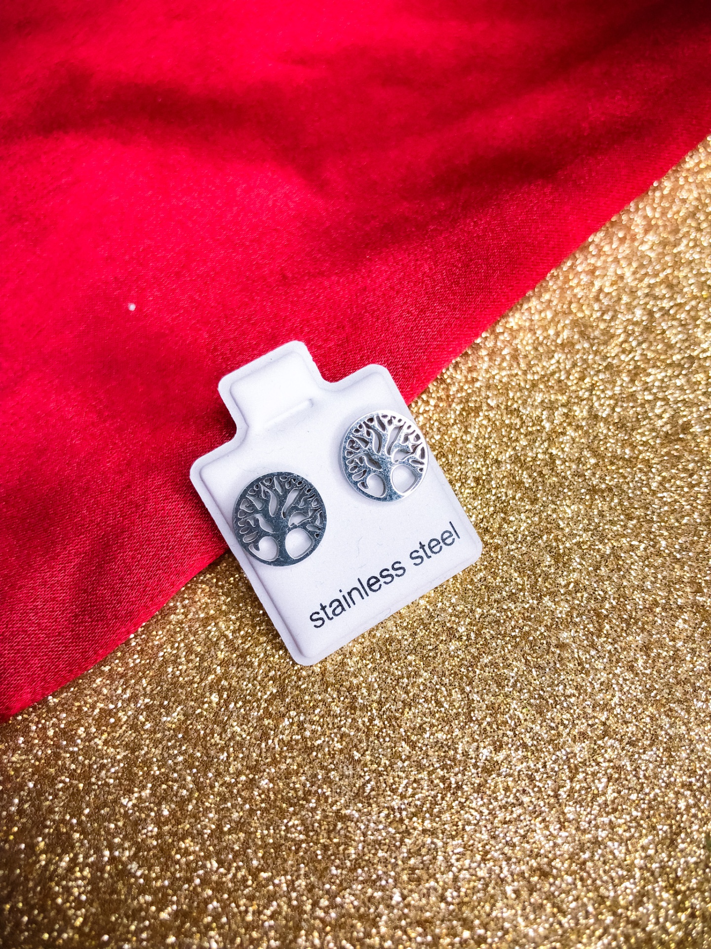 Brincos de aço inoxidável com design de árvore em cartão branco sobre fundo dourado e vermelho.