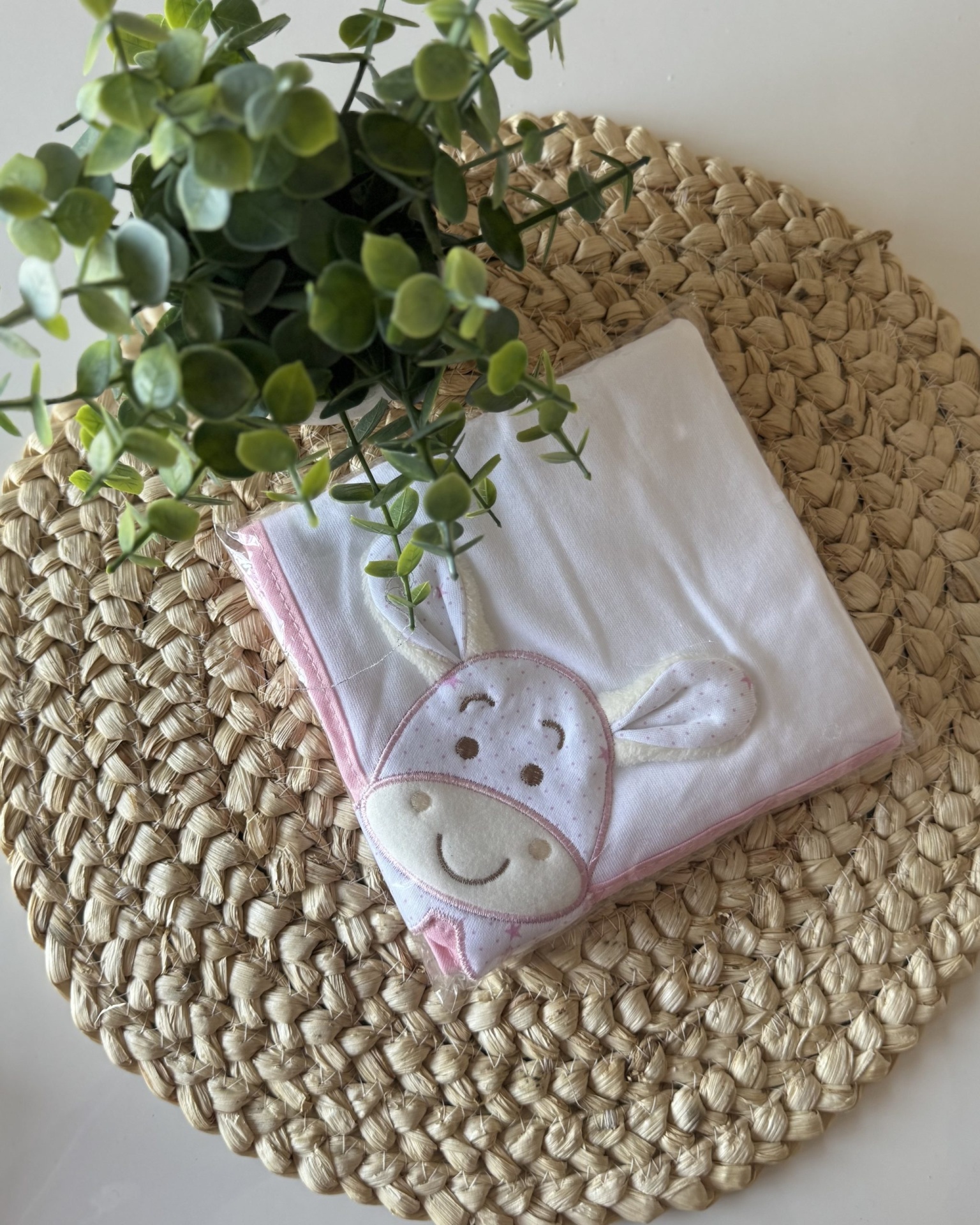 Manta branca com bordas rosa e desenho de crocodilo, sobre suporte redondo de fibra trançada e planta verde